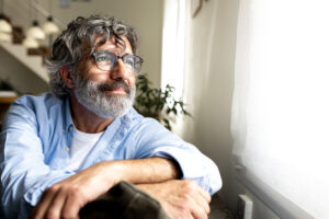 Thoughtful mature man with eyeglasses and gray hair looking outside the window while sitting at home