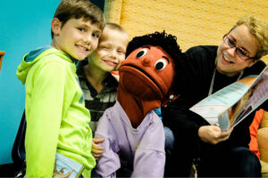 Two kids pose for a picture with a puppet while a woman reads a book to them.