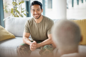 Man in combat fatigues sits on couch talking with someone whose back is to the camera