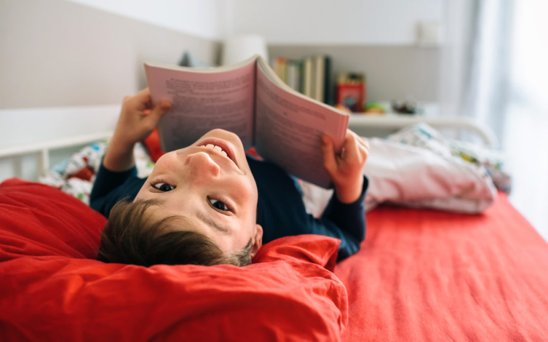 Boy looking back to the camera while reading a book lying on the bed