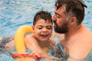 disabled boy with a float laughs as he plays and swims in a pool with his bearded father. 