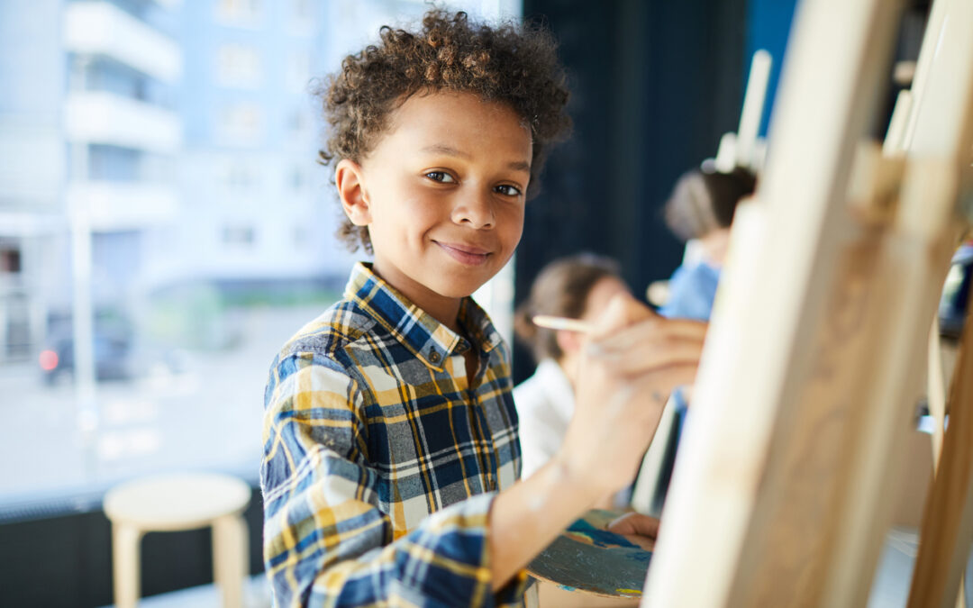 Happy mixed-race pupil looking at camera while standing by easel and painting at lesson in studio of arts