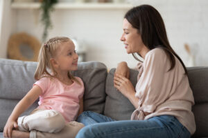 Happy young mother and little preschooler daughter sit on couch in living room talking and chatting, smiling small girl child enjoy leisure time with mom speak communicate spend time together