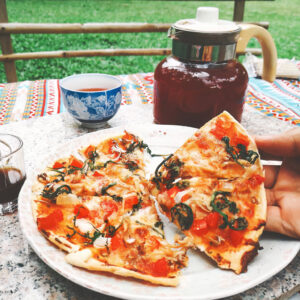 A hand holds a slice of pizza with tomatoes and basil, with a plate of pizza, tea in a cup and glass, and a pitcher on a colorful tablecloth outdoors.