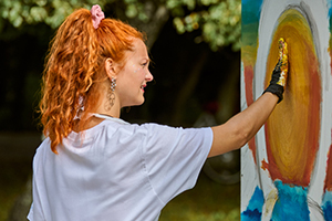 Young woman with curly red hair painting bright yellow sun on large canvas
