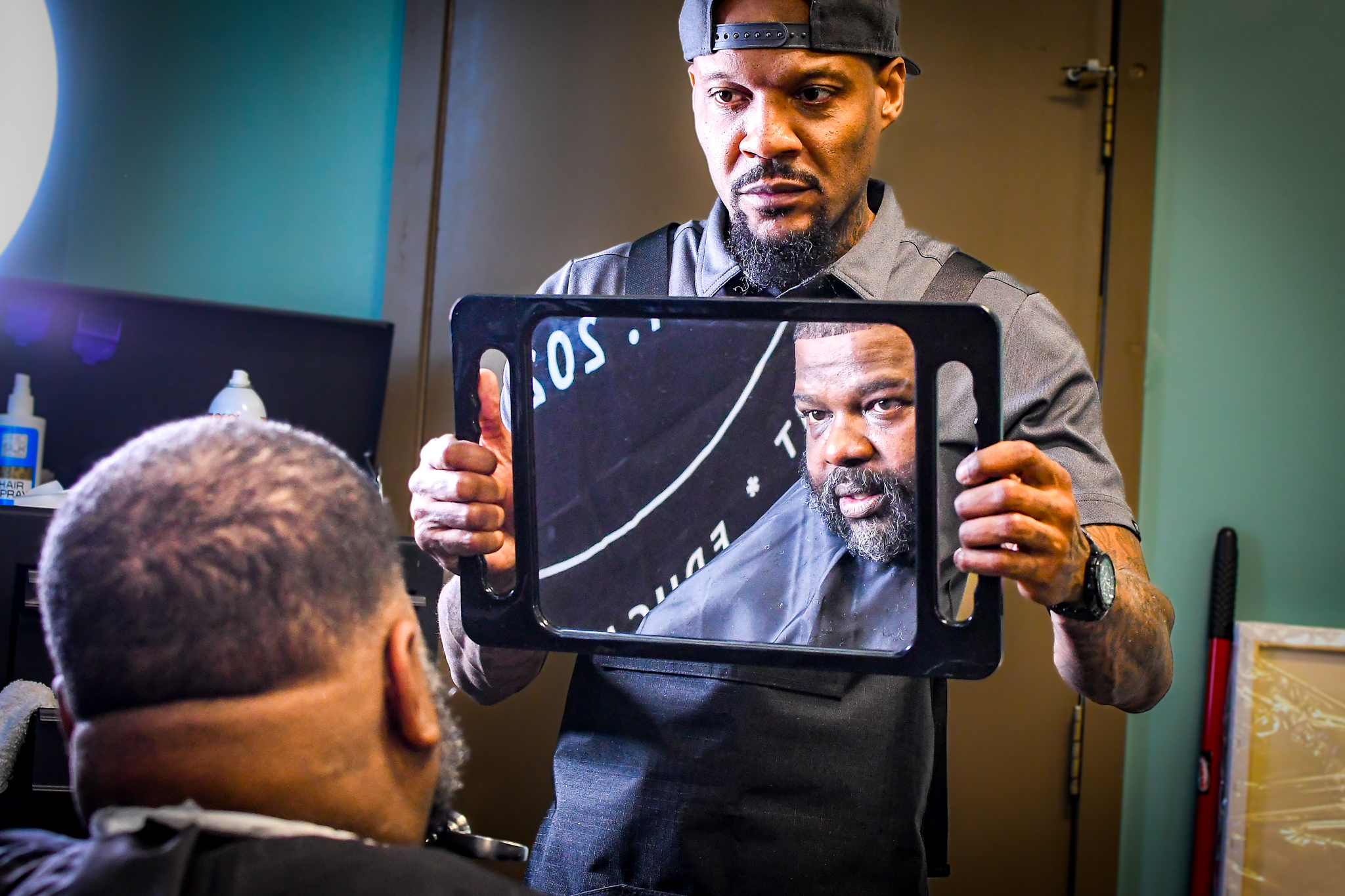 An African American man holds a mirror showing another African American man his haircut
