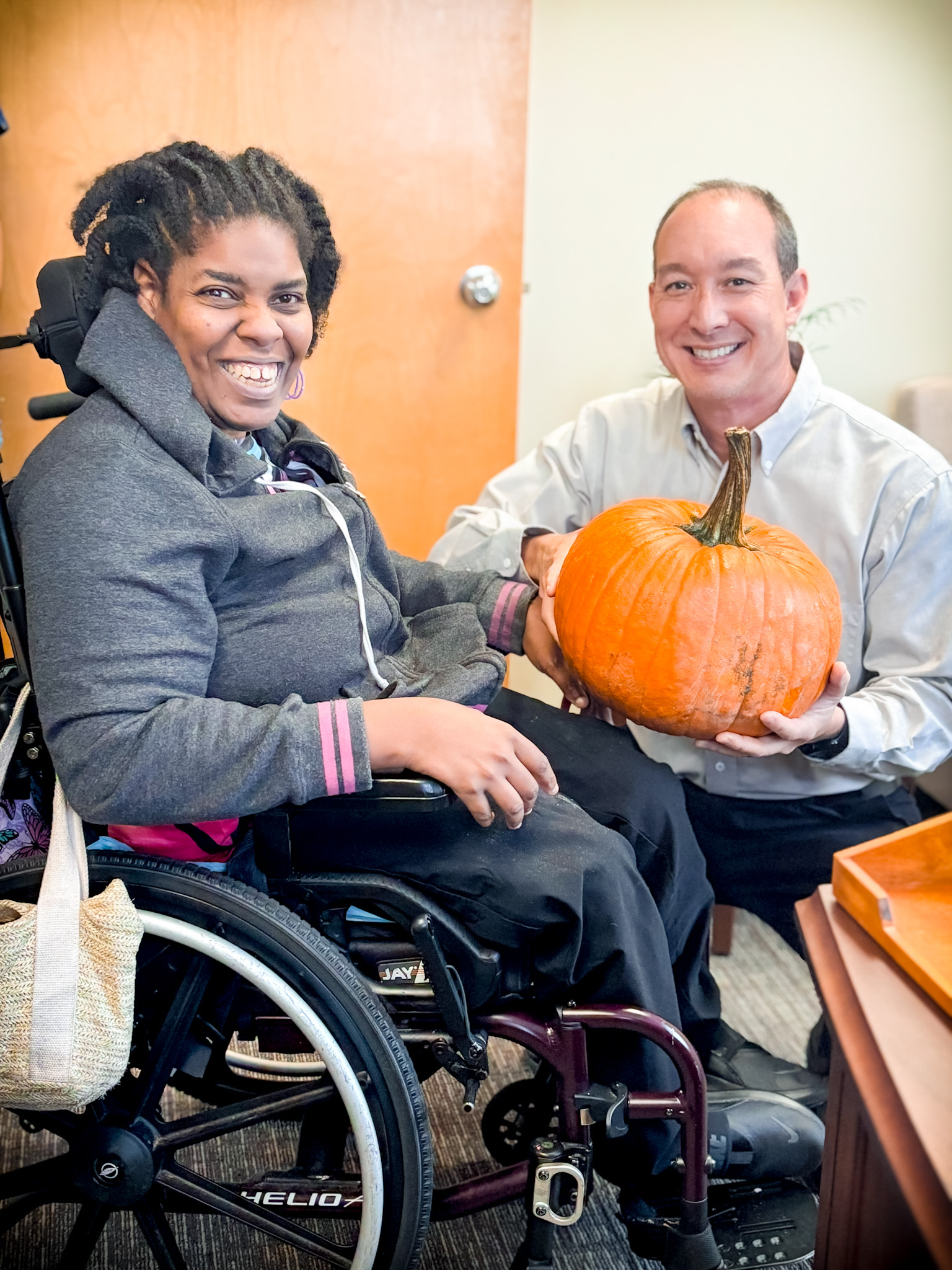 A balding man holding a pumpkin kneels next to an African American young woman in a wheelchair