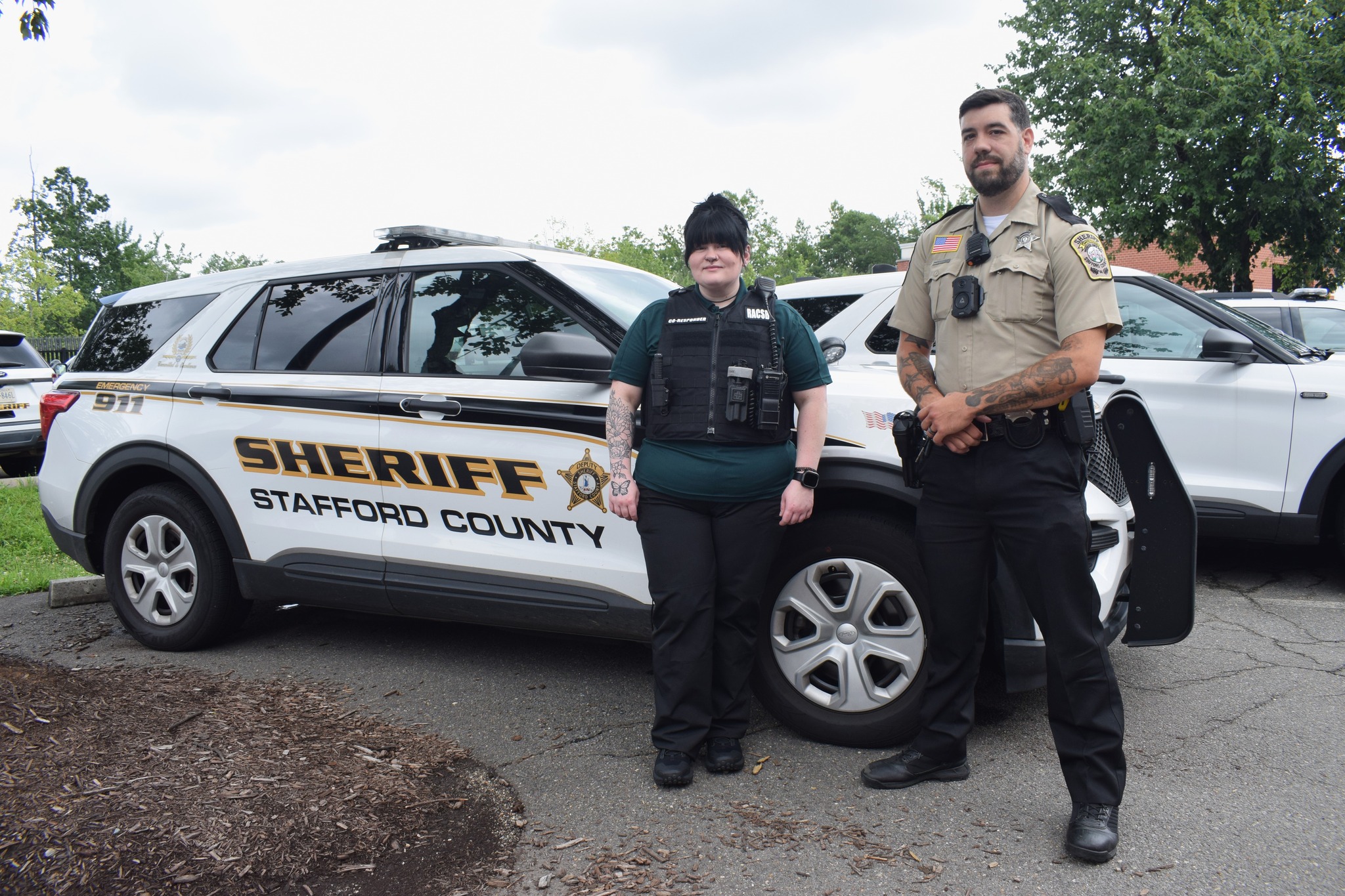 A woman stands next to a man wearing a deputy uniform in front of a police car that reads "Sheriff Stafford County"