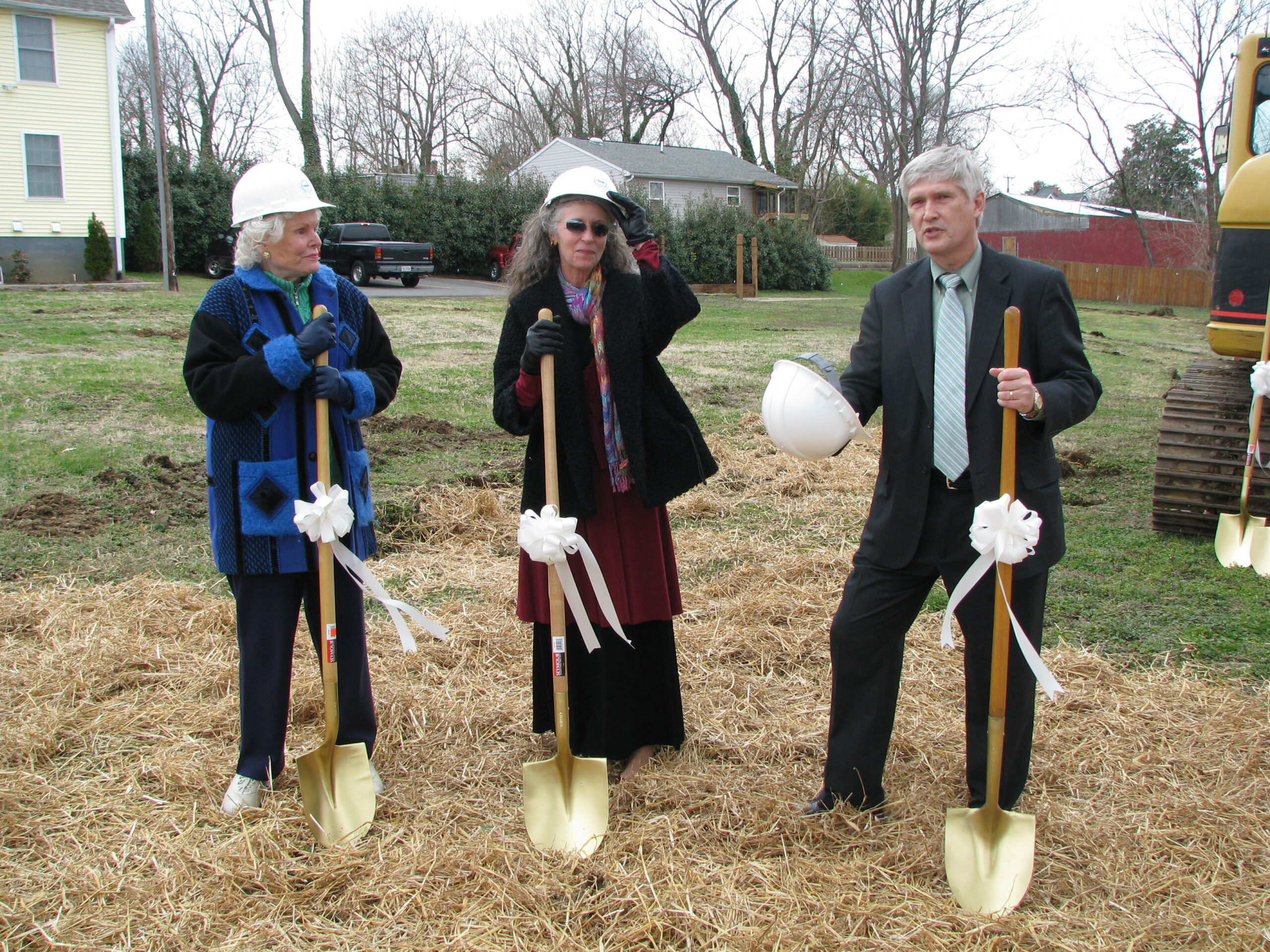 Two women wearing hardhats and holding shovels listen to a man speaking who is holding a hardhat in one hand and a shovel in the other