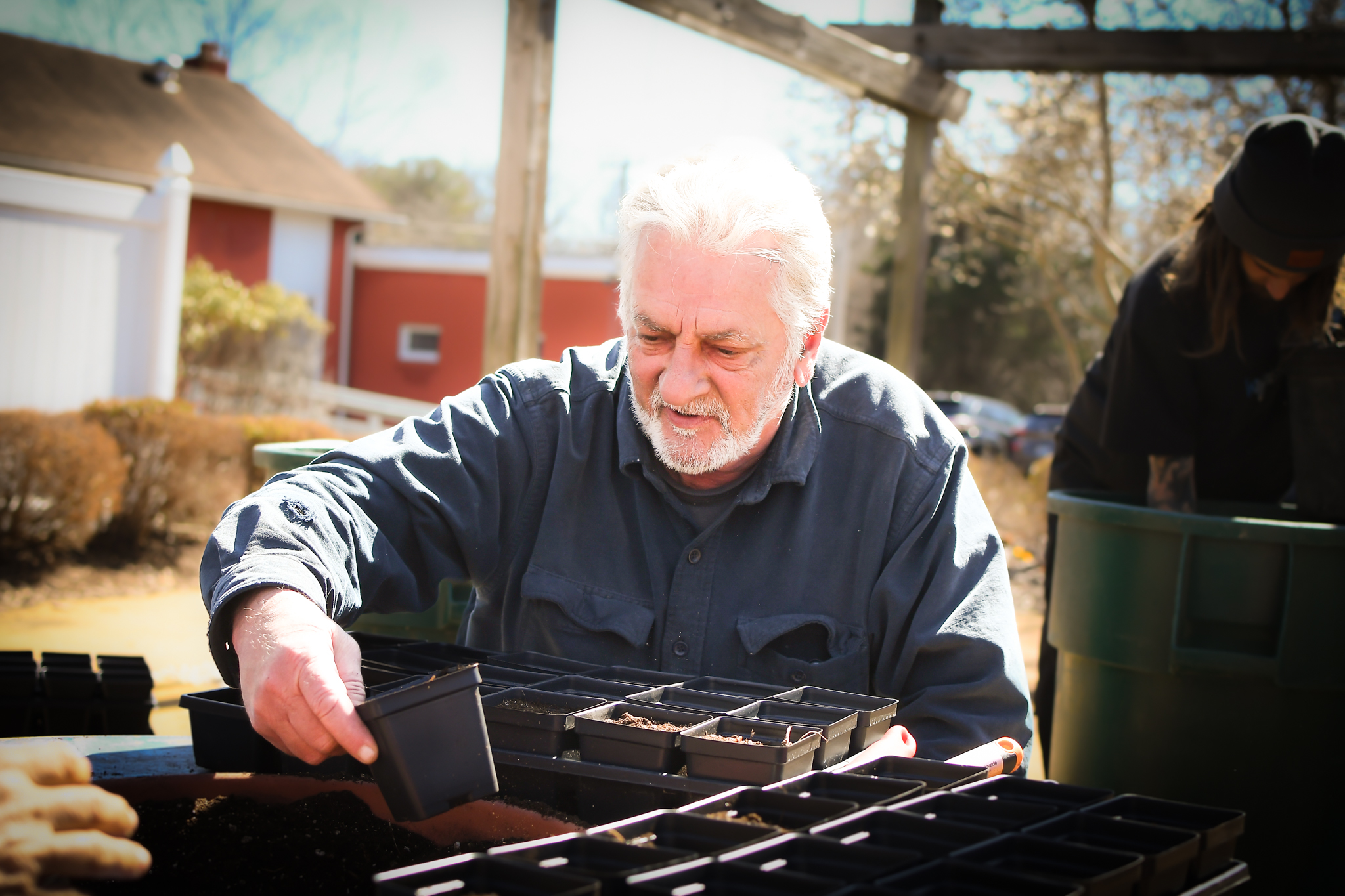 A white man with gray hair and a beard plants seeds in a tray 