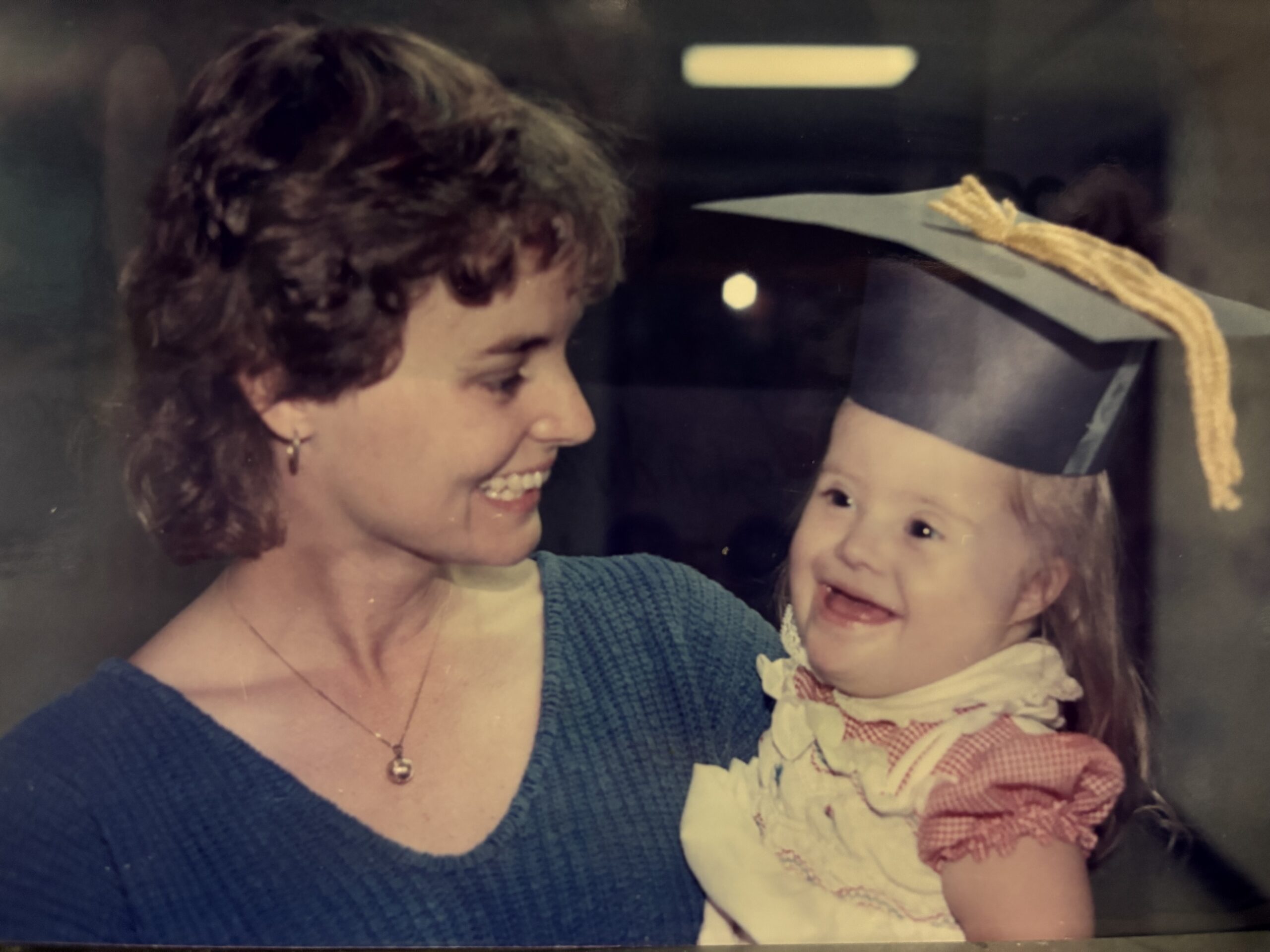 A smiling woman holds a toddler with Down syndrome who is wearing a paper graduation hat