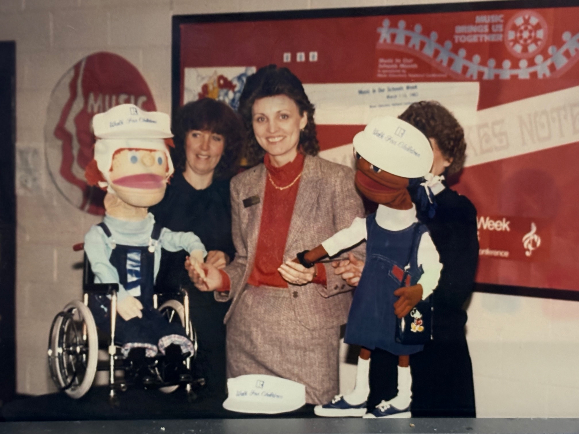 A woman in a business suit stands between two woman holding large puppets