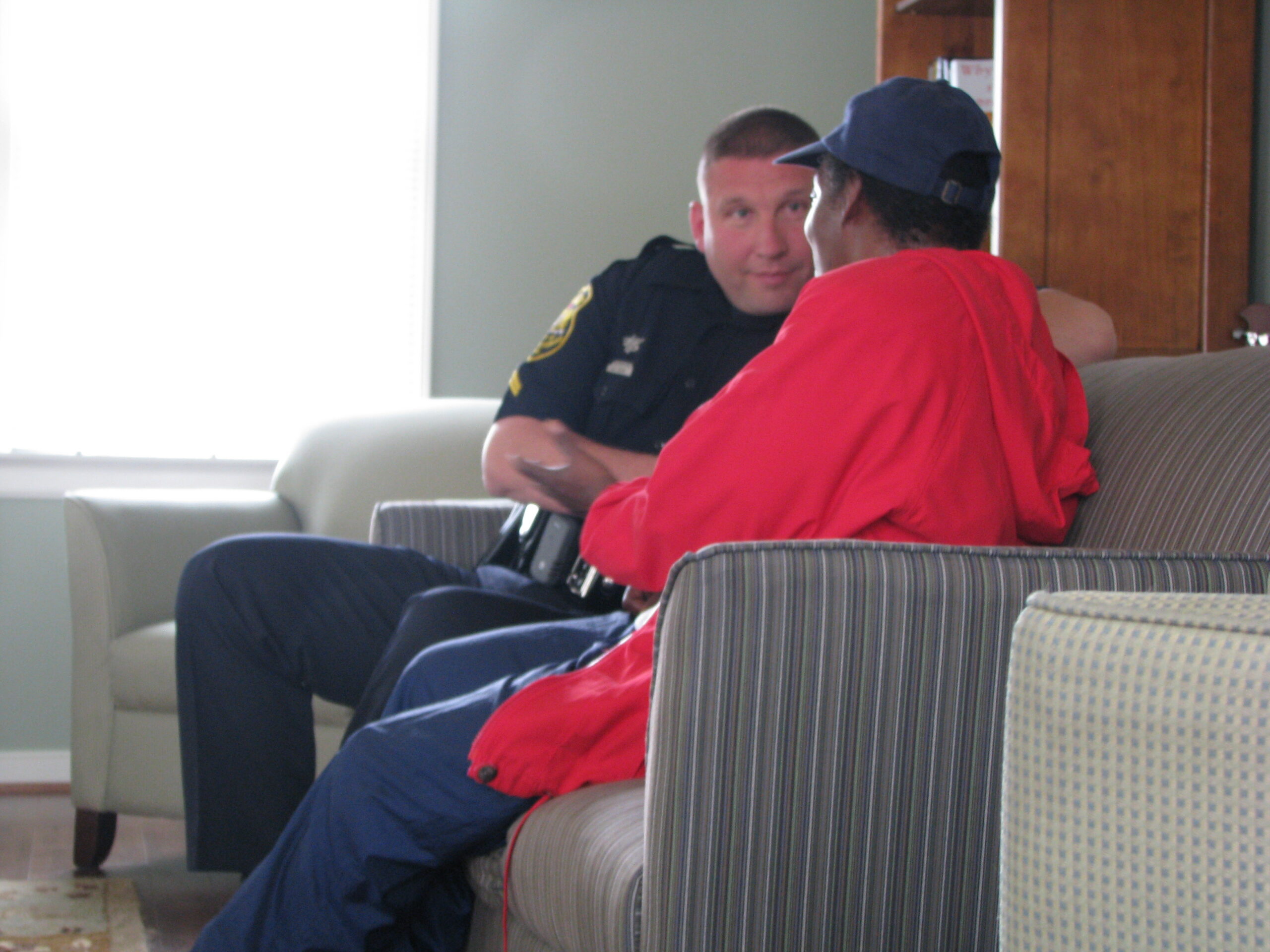 A police officer talks to a young adult on a couch with a bookcase behind them
