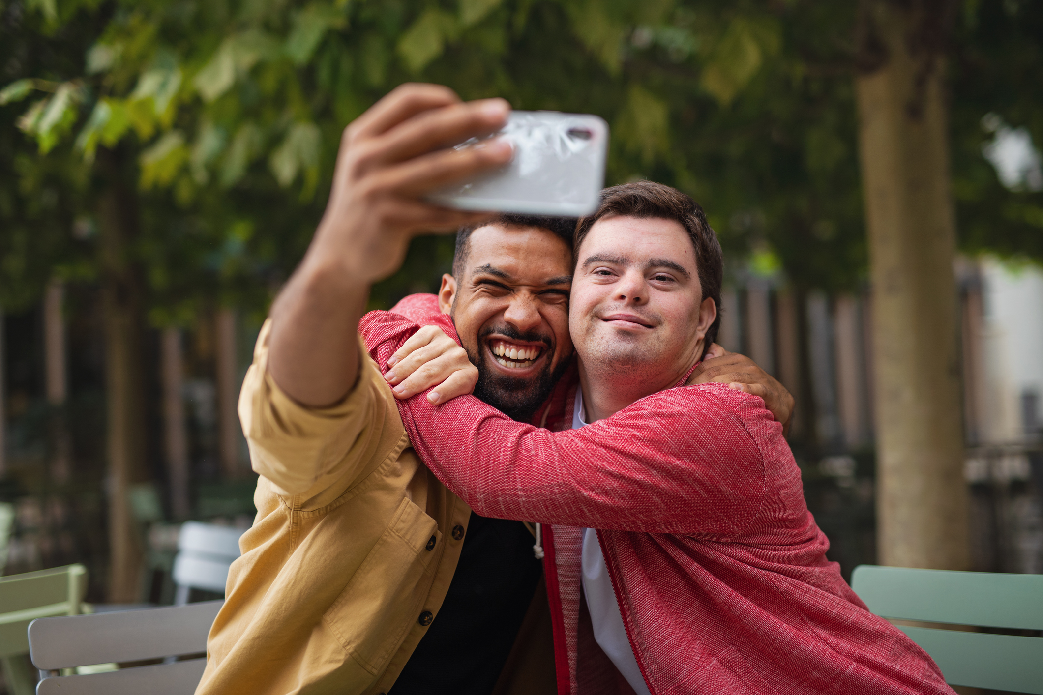 A young man with Down syndrome and a young man in a jacket hold a smartphone and take a selfie outside